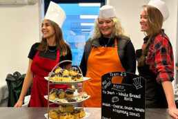 A photo of three women smiling behind a stand of baked goods and a menu board.