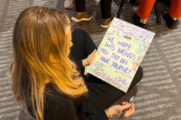 A photo showing a woman holding a sign that says 'We hope this brings you joy on your journey'.