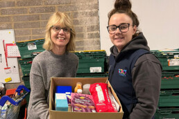 A picture displaying two woman holding a cardboard box full of packaged food items.