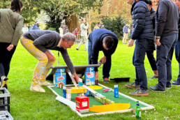 A picture showing two men bending down to help build a mini golf hole out of items such as cereal and tea bags