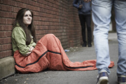 An image of a homeless woman sat in an orange sleeping bag on the floor