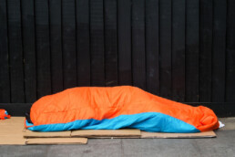 An image showing someone completely covered by a sleeping bag lying on the floor on cardboard