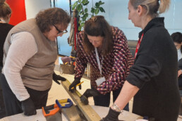 A picture showing 3 woman with safety gloves on measuring out a piece of wood.